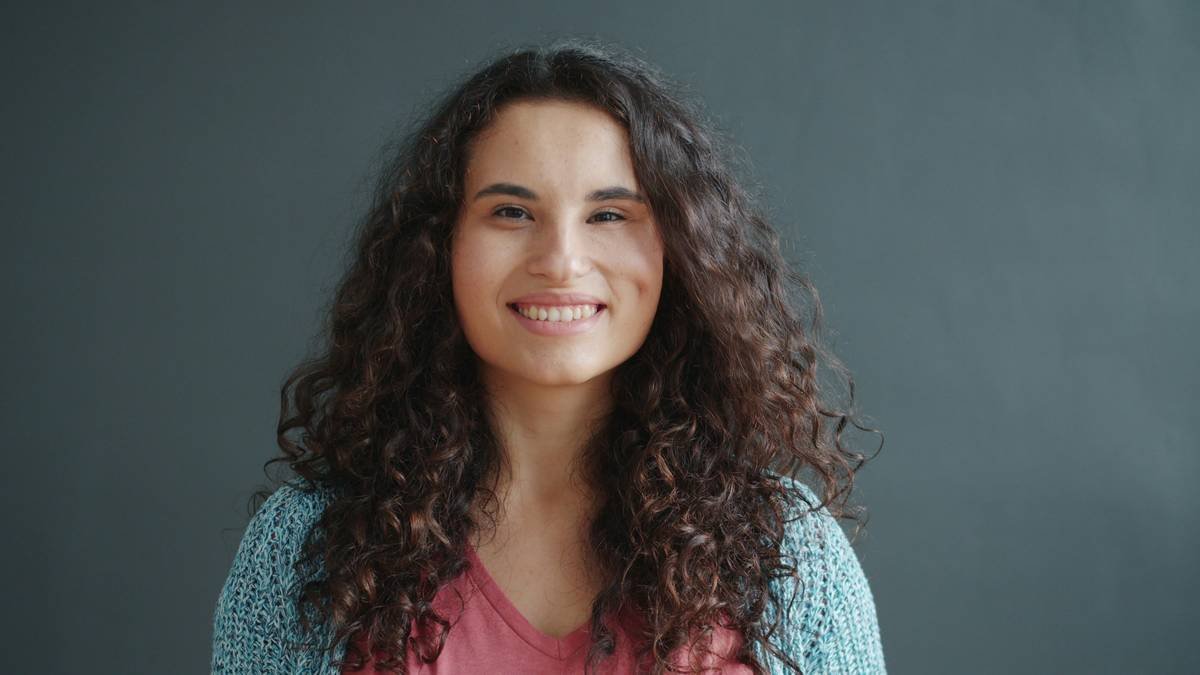 Cute lady is showing OK hand gesture standing indoors on dark background with joyful smile, good-looking young woman is wearing casual clothing.